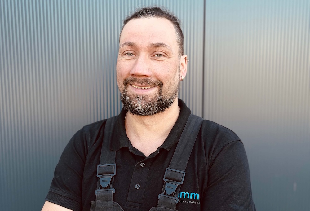 Lächelnder Handwerker in schwarzem Poloshirt mit Arbeitslatzhosen, vor einer modernen, strukturierten Hintergrundwand.